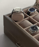 Three wristwatches are nestled in a TAWBURY Bayswater Watch Box with beige 55mm pillows. One silver watch is highlighted on a raised section of the elegant brown leather case.